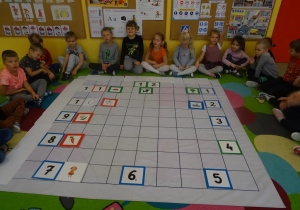 Grupa dzieci siedzi wokół rozłożonej na dywanie macie do kodowania. Na macie ułożony jest zegar z tabliczek z cyframi oraz obrazki, które określały co dzieci robią o danej godzinie.